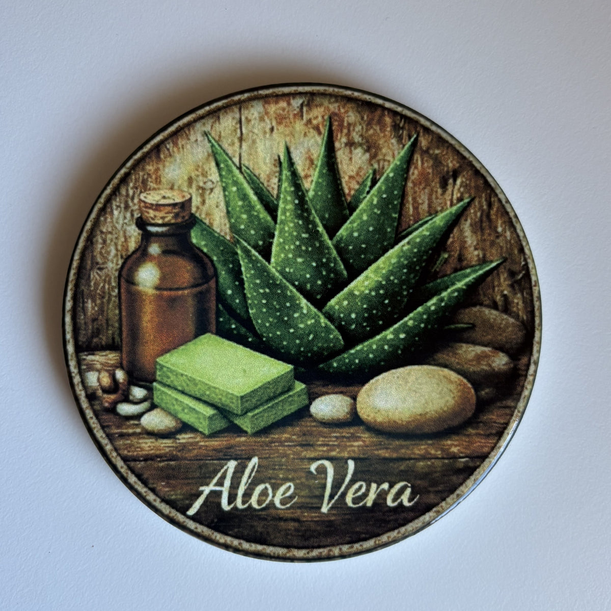 Round coaster featuring an illustrated aloe vera design with green aloe leaves, a brown bottle, green soap bars, stones, and the words 'Aloe Vera' on a dark background.