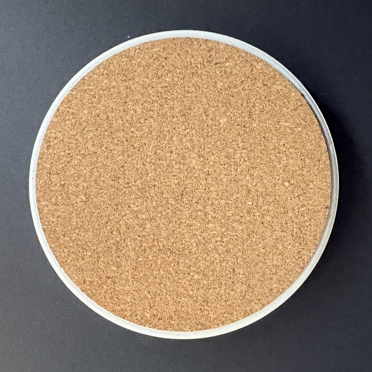 A round cork coaster with a white rim, photographed from directly above on a dark gray background.