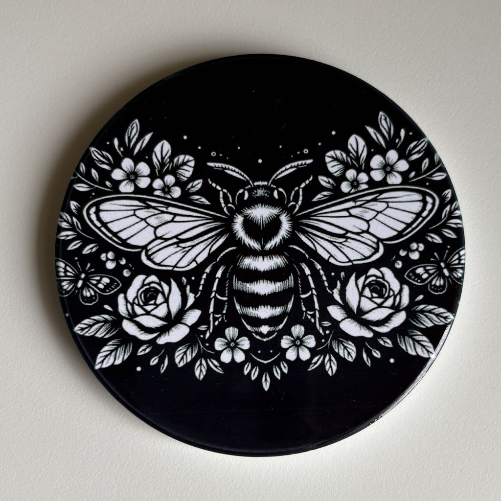 A round black coaster featuring a detailed white graphic illustration of a bumblebee surrounded by flowers, displayed on a neutral off-white background.