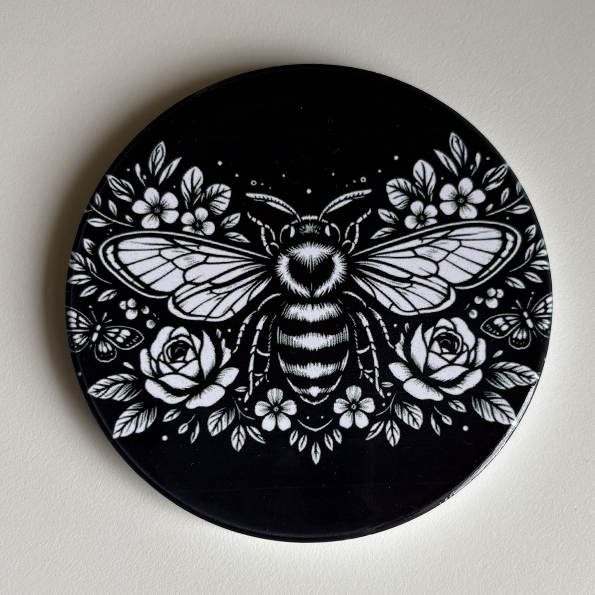 A round black coaster featuring a detailed white graphic illustration of a bumblebee surrounded by flowers, displayed on a neutral off-white background.