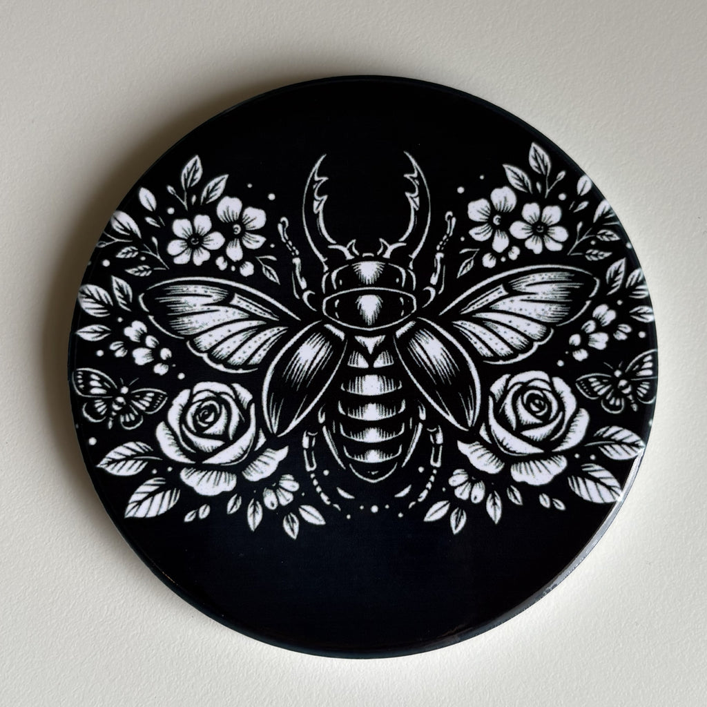 A round black coaster featuring a detailed white line art illustration of a beetle, roses, and flowers, displayed against a light off-white background.