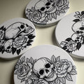 Four coasters with black and white skull and floral designs on a dark background