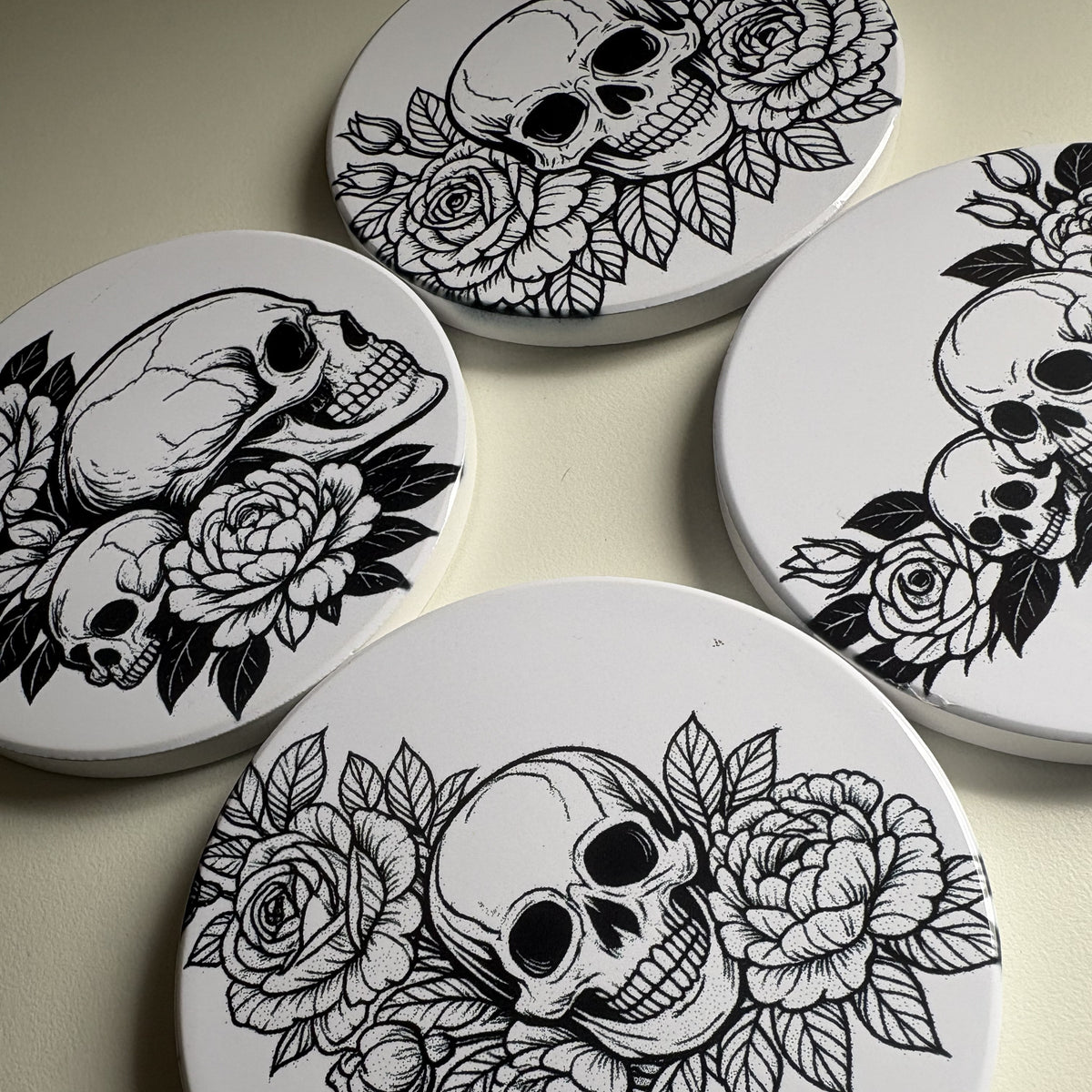 Four coasters with black and white skull and floral designs on a dark background