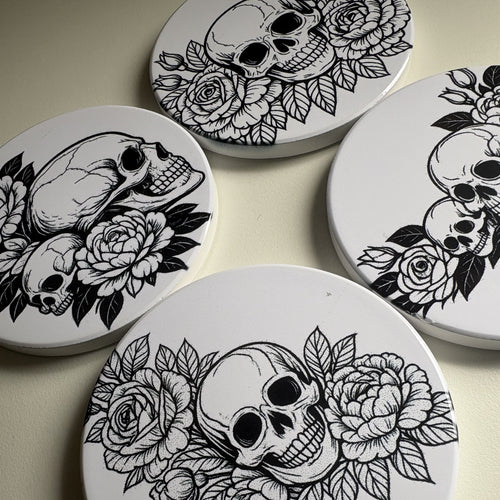 Four coasters with black and white skull and floral designs on a dark background