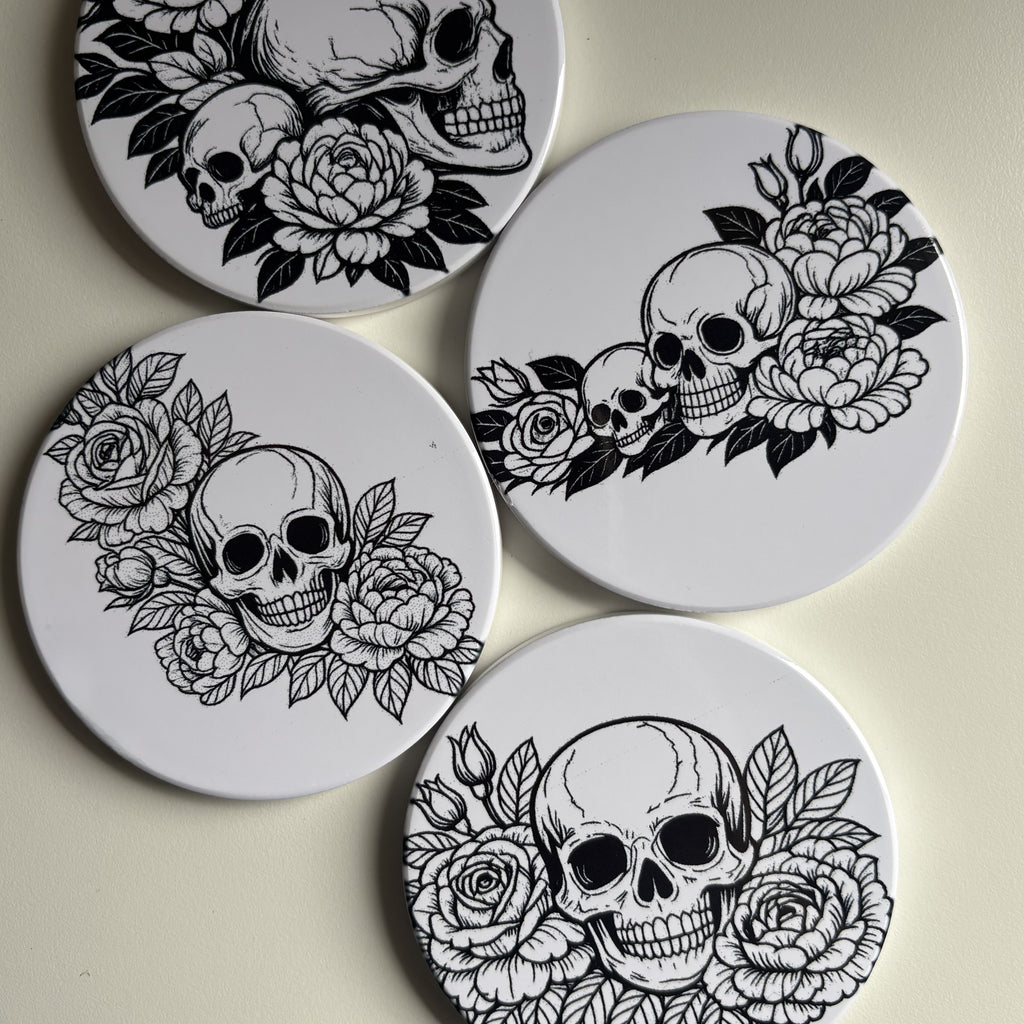 Four round white coasters featuring black ink illustrations of skulls surrounded by detailed roses and leaves, arranged on a light beige surface.