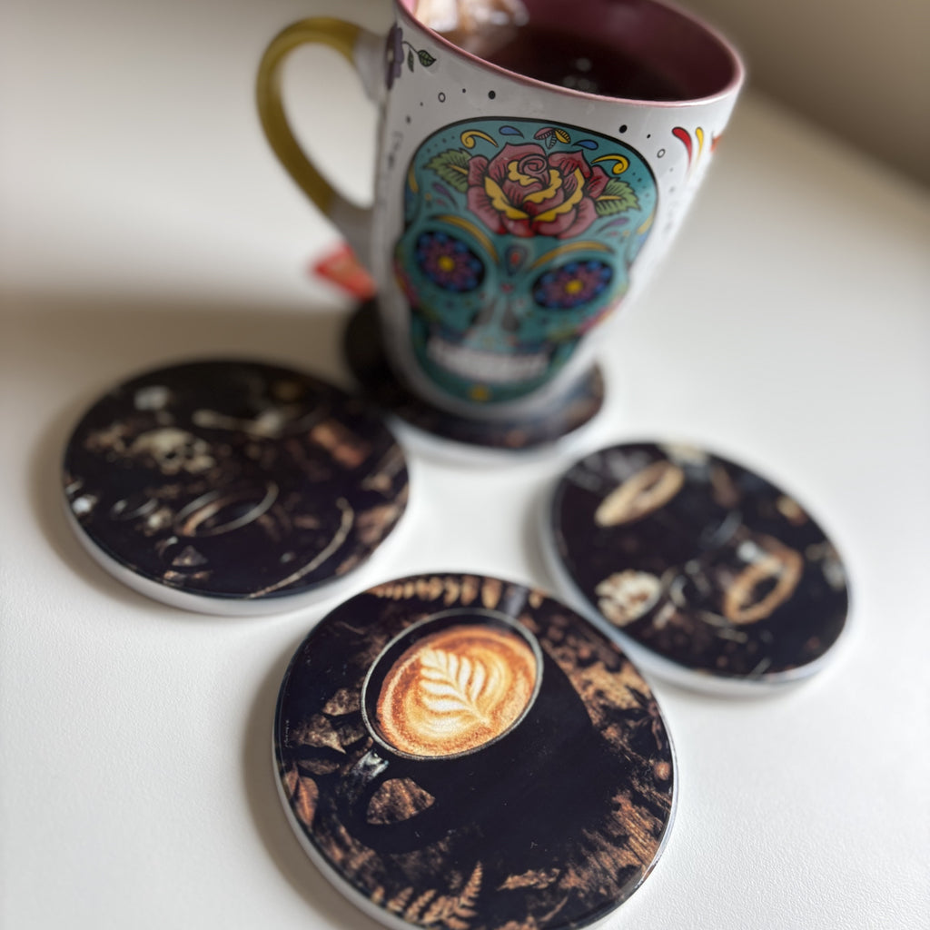 A white ceramic mug with a colorful sugar skull design sits on a coaster, with three matching round coasters displaying coffee-themed prints on a light surface.