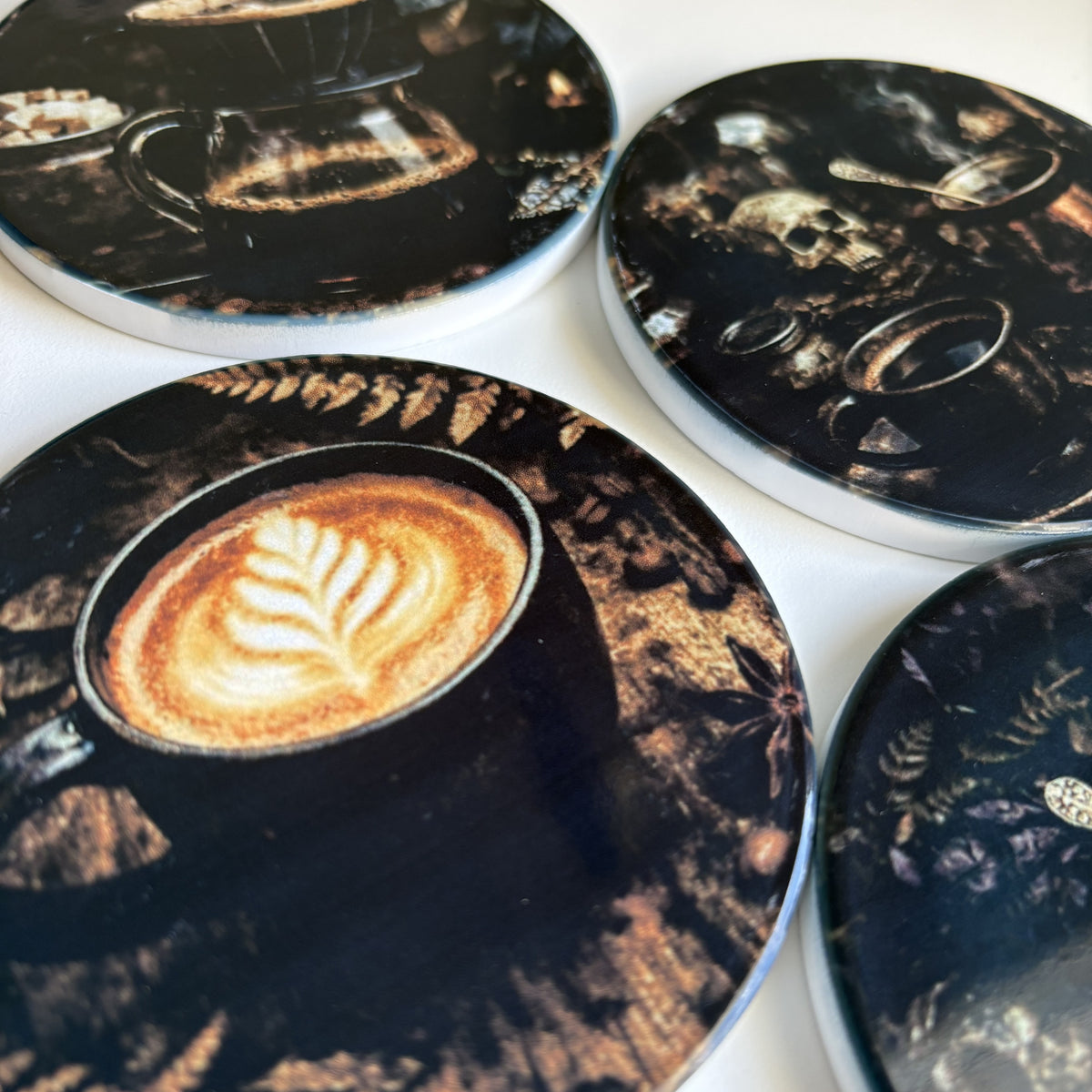 A set of round coasters featuring dark, moody coffee-themed illustrations, including latte art, espresso cups, skulls, and botanical details on a white surface.