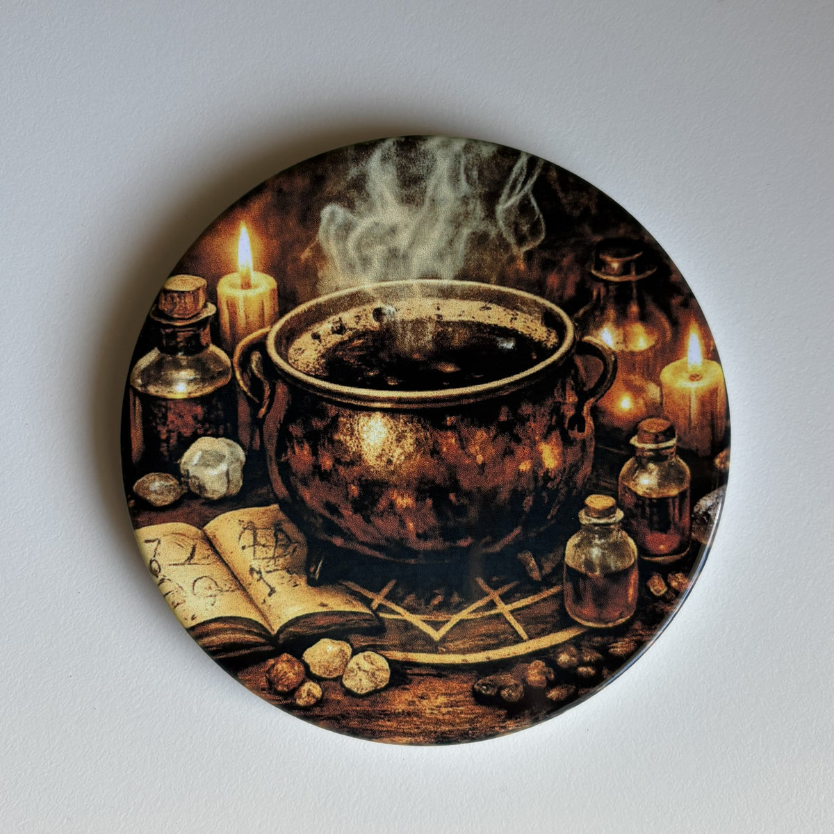 A coaster featuring a dark, mystical illustration of an alchemy cauldron with steaming smoke, surrounded by lit candles, small glass bottles, an open book with symbols, and stones, photographed against a plain light gray background.