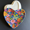 A heart-shaped ceramic dish with a vibrant multicolor floral pattern on a dark blue background, shown with its off-white lid behind it on a dark gray surface.