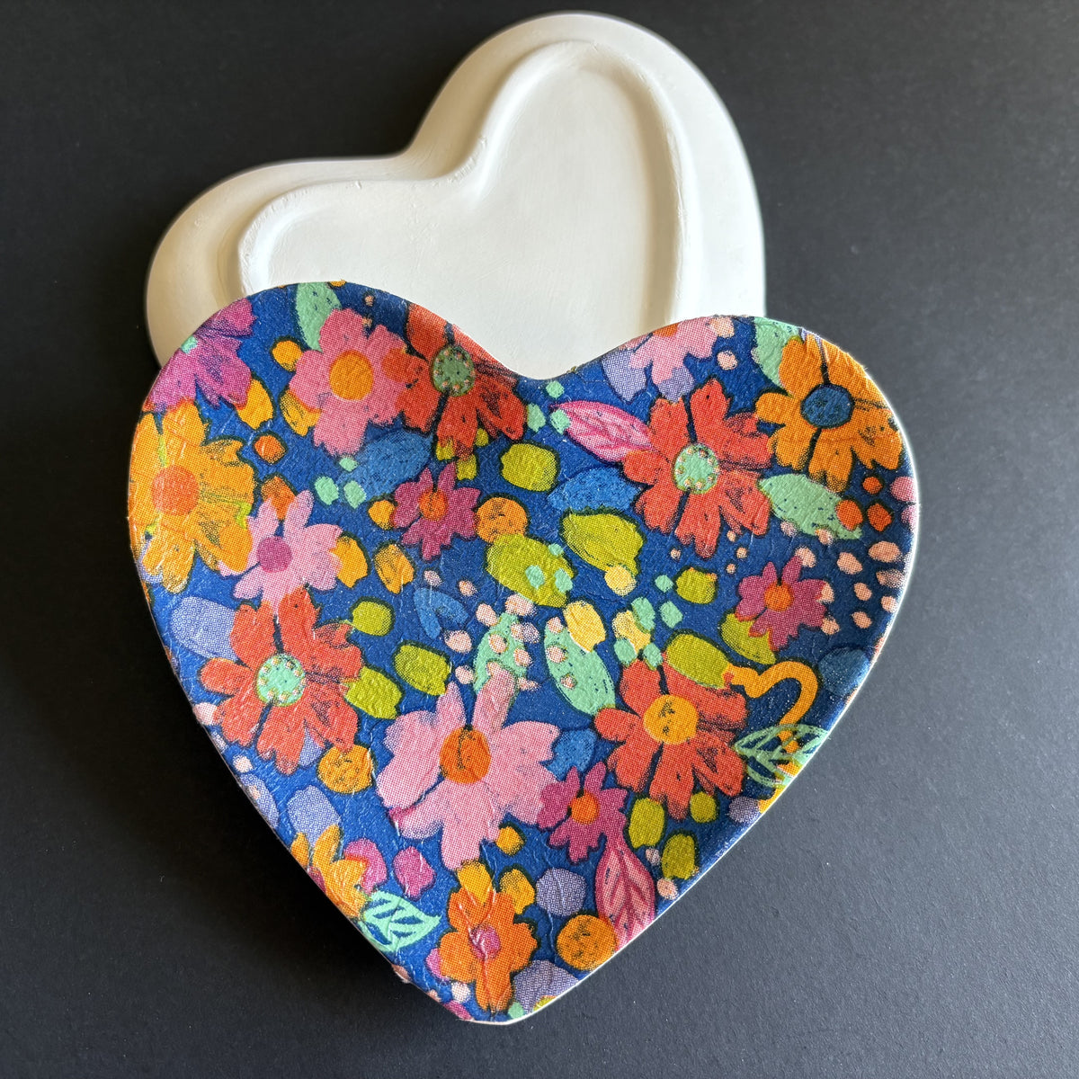 A heart-shaped ceramic dish with a vibrant multicolor floral pattern on a dark blue background, shown with its off-white lid behind it on a dark gray surface.