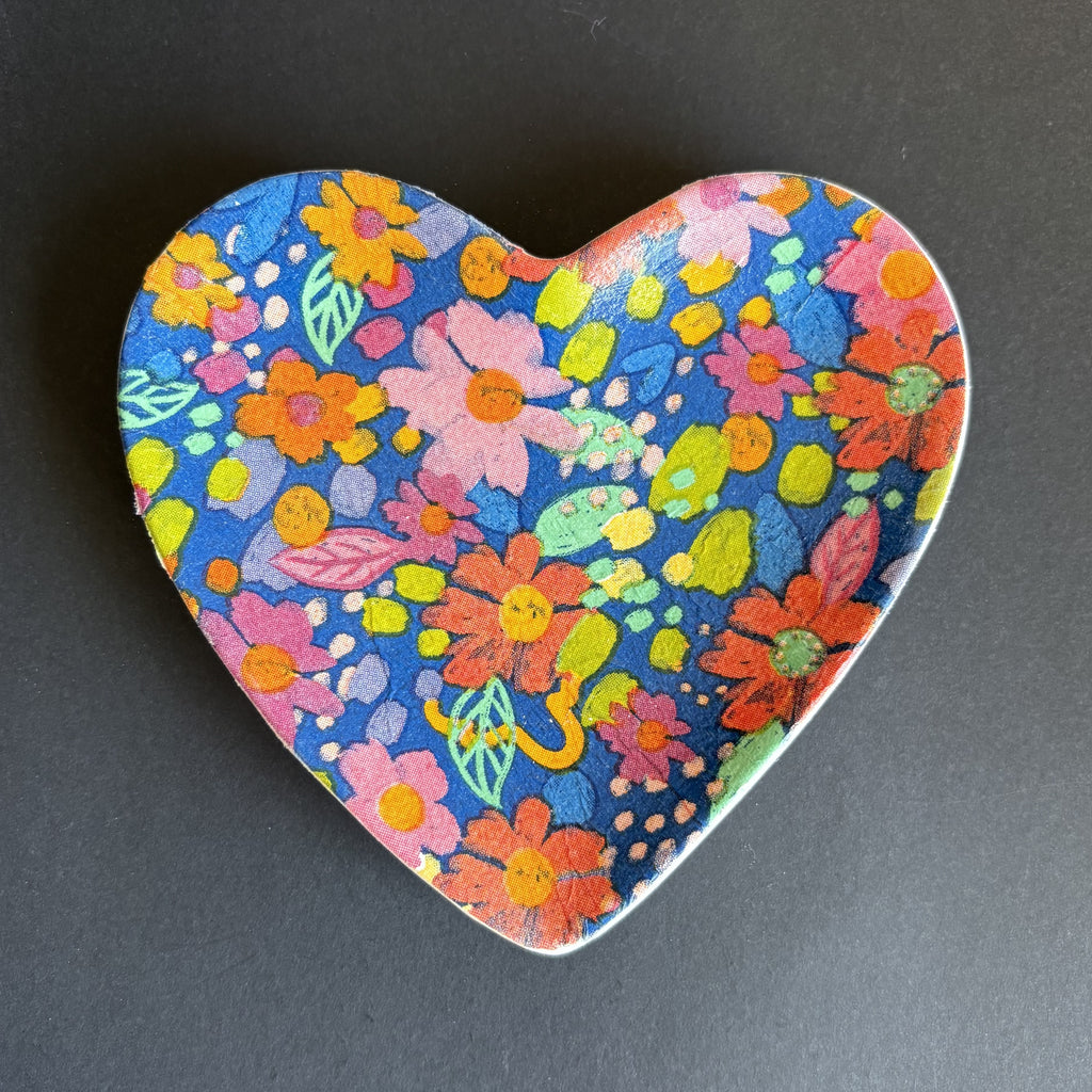 A heart-shaped ceramic dish featuring a vibrant, hand-painted style multicolor floral pattern on a dark blue background, set against a dark gray surface.
