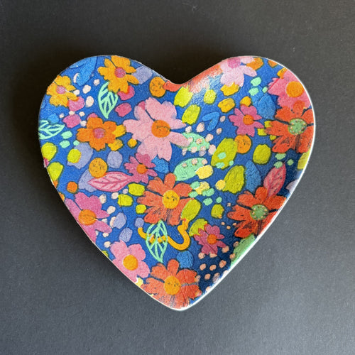 A heart-shaped ceramic dish featuring a vibrant, hand-painted style multicolor floral pattern on a dark blue background, set against a dark gray surface.