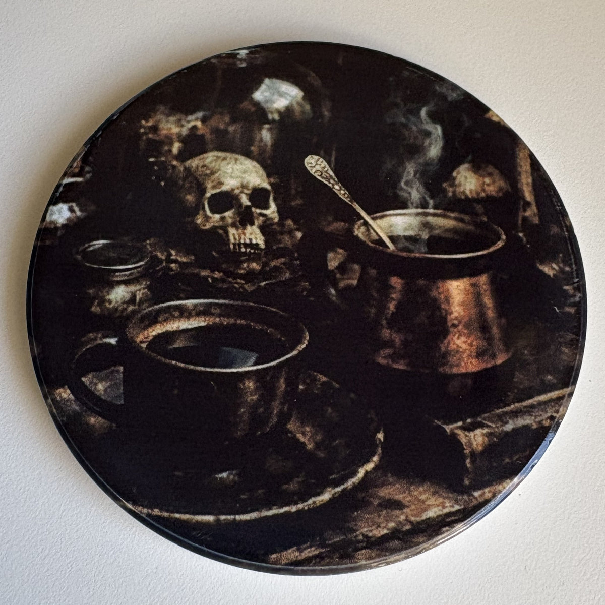 A round coaster featuring a dark, gothic illustration of a skull, a steaming mug, and a copper pot on a black background, shown against a plain off-white wall.