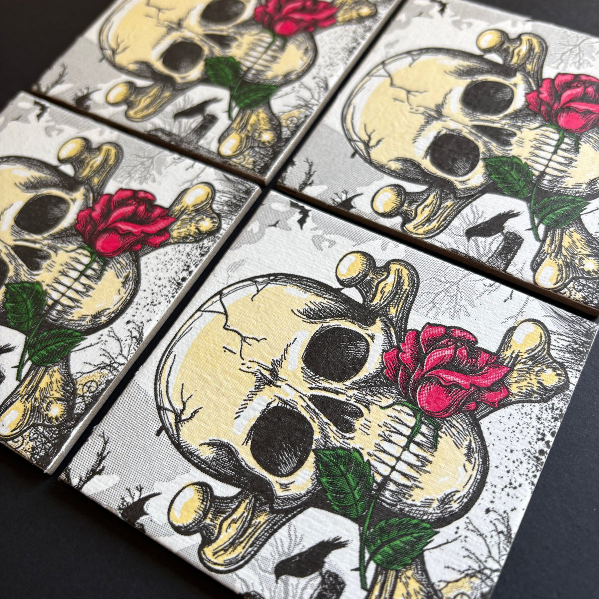 Four square magnet arranged in a grid, each printed with a skull over crossed bones, black eye sockets, and a red rose with green leaves on a dark background.