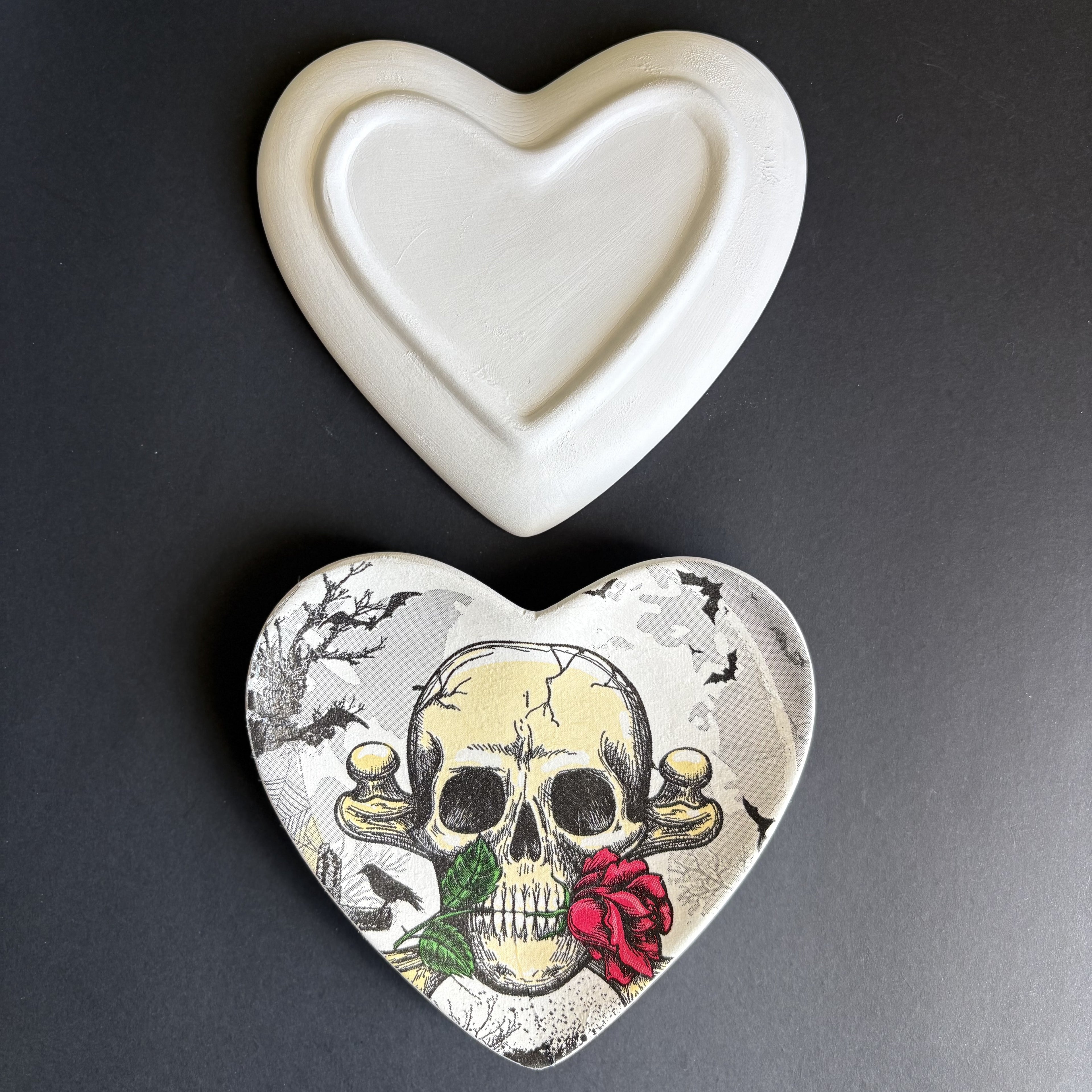 A heart-shaped ceramic dish with a white glazed finish on the top half and a detailed skull and rose illustration on the bottom half, shown on a dark gray background.