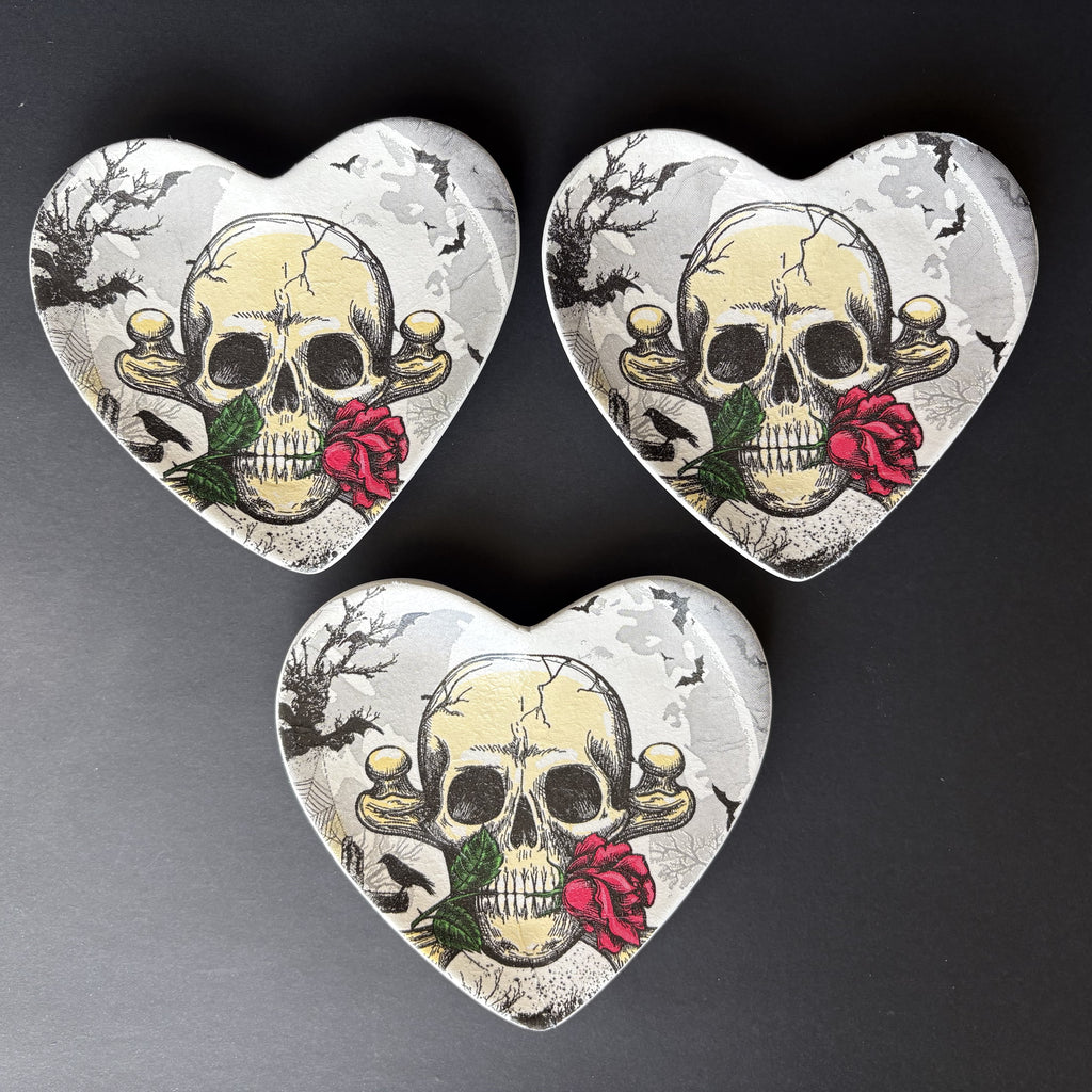 Three heart-shaped trinket tray with a vintage skull and red rose design, arranged on a dark gray background.