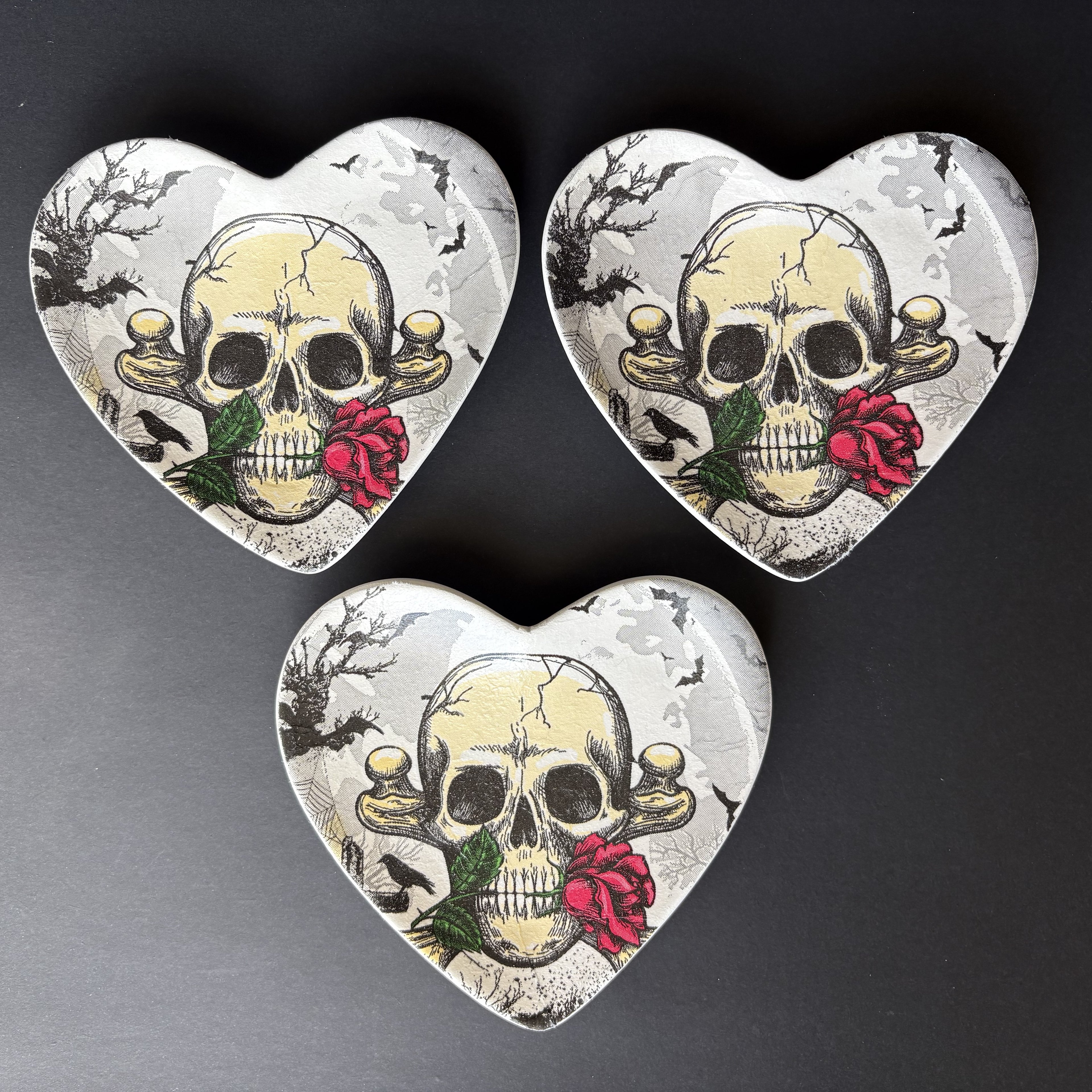 Three heart-shaped trinket tray with a vintage skull and red rose design, arranged on a dark gray background.