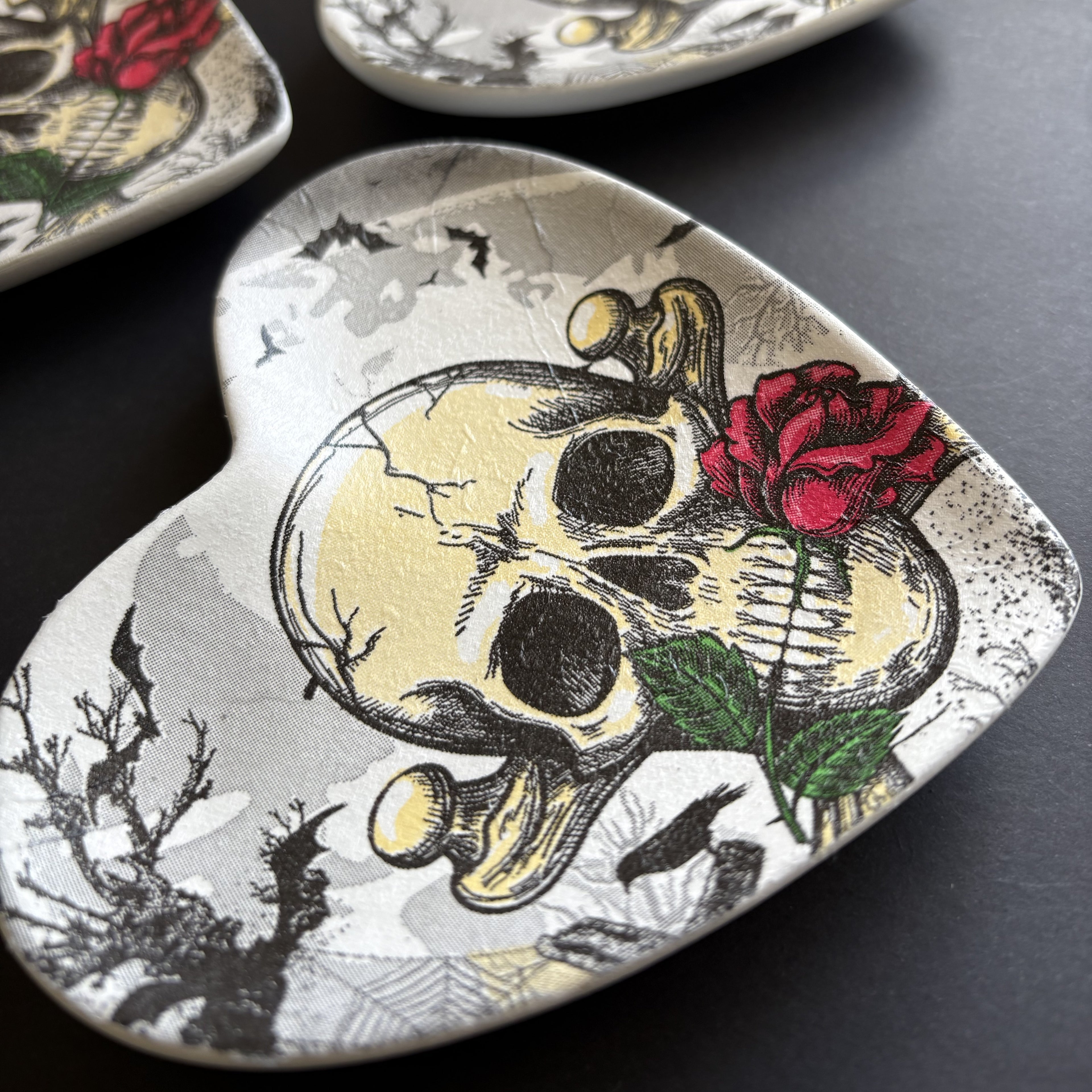 A heart-shaped ceramic trinket tray or dish featuring a detailed illustration of a skull with a red rose, set against a dark textured background.