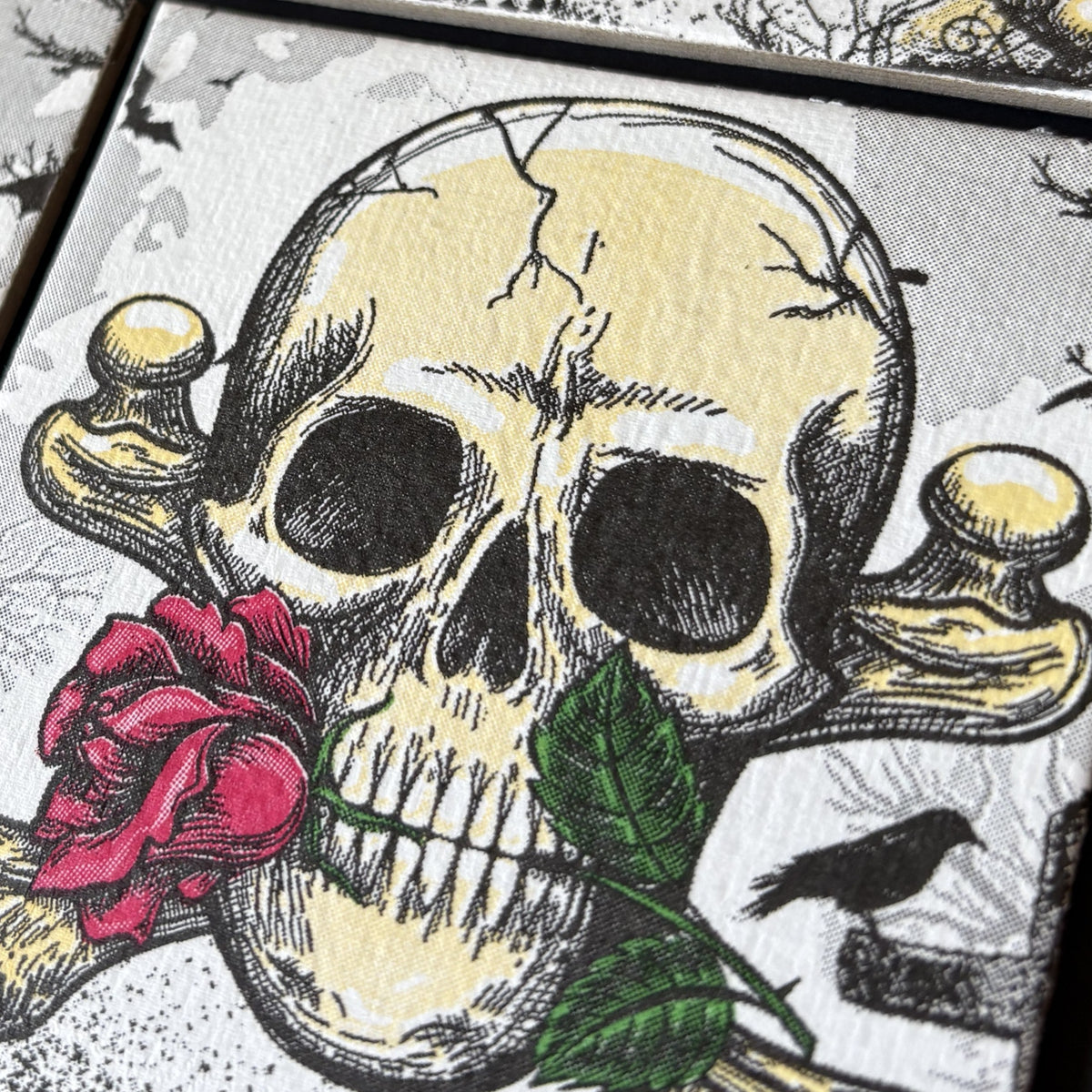 Close-up of a square art print featuring a stylized skull and crossbones with a red rose and green leaf, on a textured off-white background with a small black bird detail.