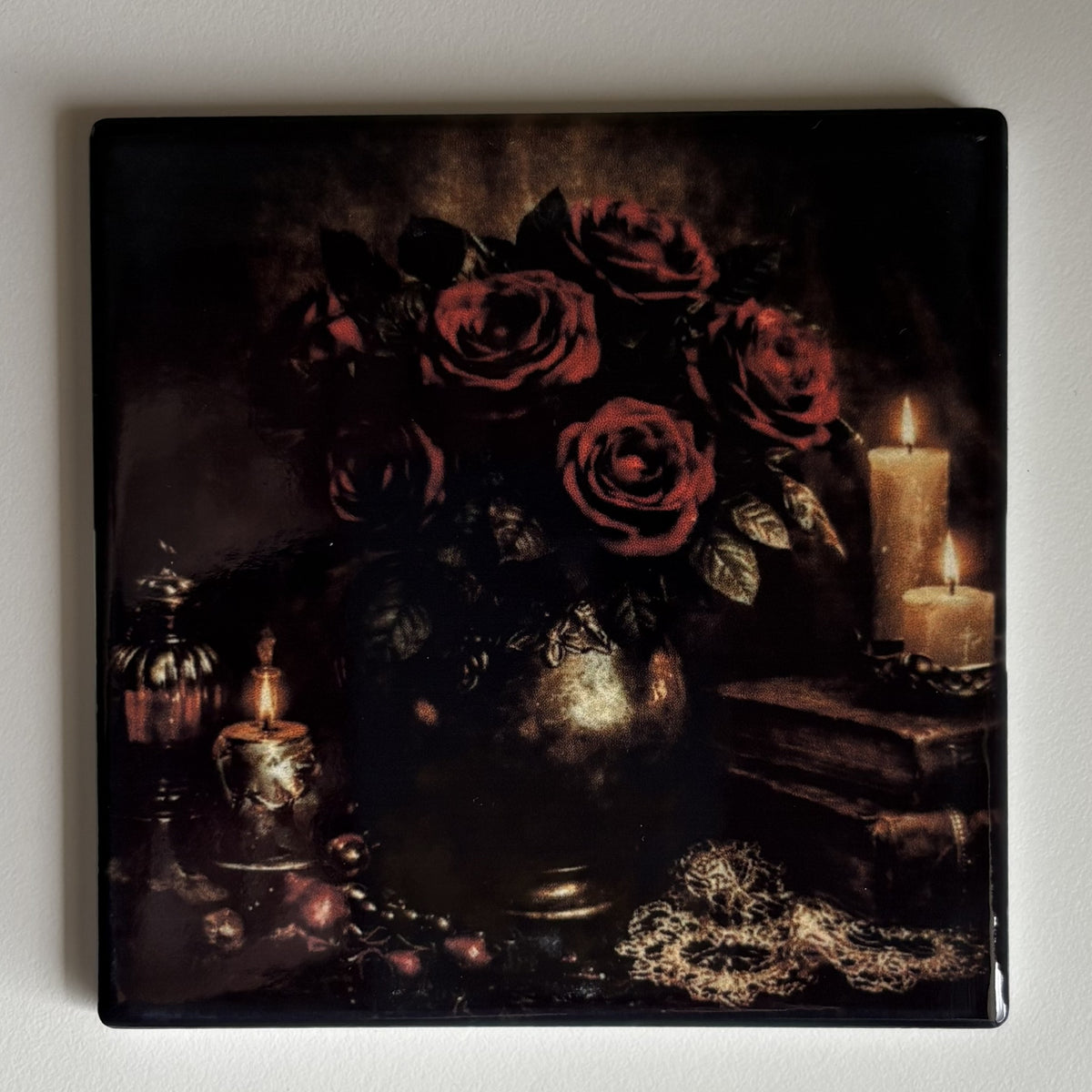 A square coaster featuring a dark gothic still life painting of red roses in a vase, surrounded by lit candles, books, and a gold chain, shown on a light gray background.