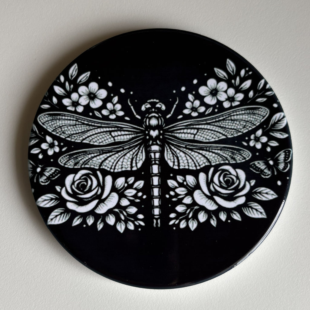 A round black coaster featuring a detailed white graphic print of a dragonfly surrounded by roses and flowers, shown on a neutral off-white background.