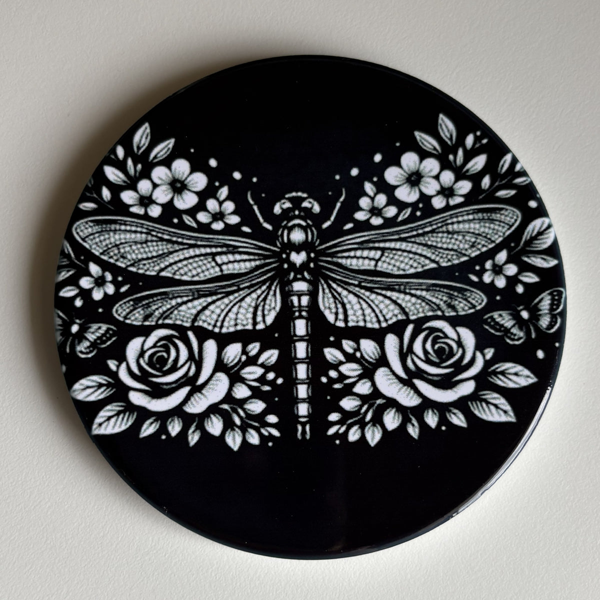 A round black coaster featuring a detailed white graphic print of a dragonfly surrounded by roses and flowers, shown on a neutral off-white background.