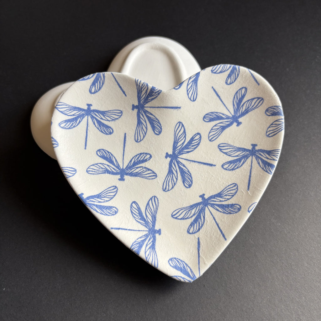 A heart-shaped ceramic dish with a blue dragonfly pattern on a cream-colored glaze, set against a dark gray background.
