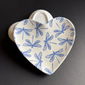 A heart-shaped ceramic dish with a blue dragonfly pattern on a cream-colored glaze, set against a dark gray background.