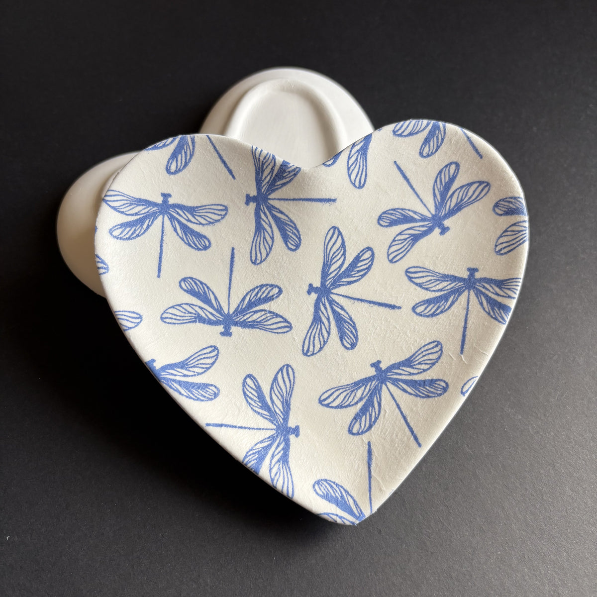 A heart-shaped ceramic dish with a blue dragonfly pattern on a cream-colored glaze, set against a dark gray background.