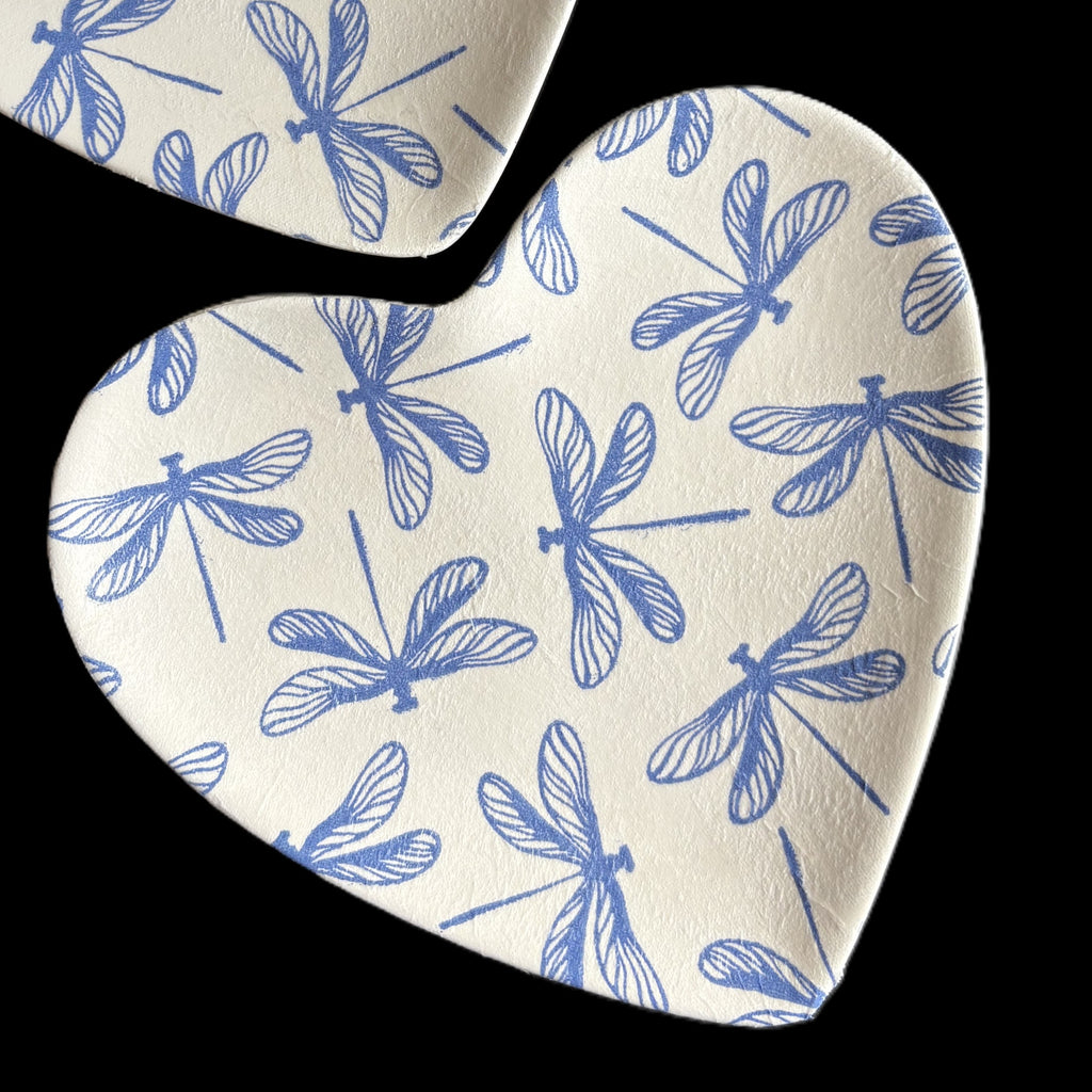 Heart-shaped ceramic TrinketTray with a blue dragonfly pattern on a white textured background, shown against a solid black backdrop.
