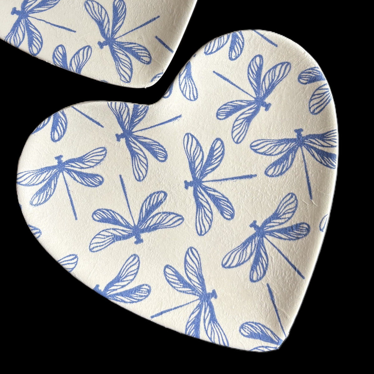 Heart-shaped ceramic TrinketTray with a blue dragonfly pattern on a white textured background, shown against a solid black backdrop.
