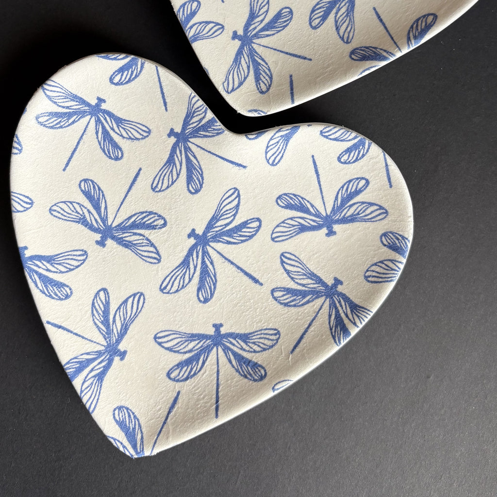 Heart-shaped ceramic Trinket Tray with a blue dragonfly pattern on a warm off-white background, shown on a dark gray surface.