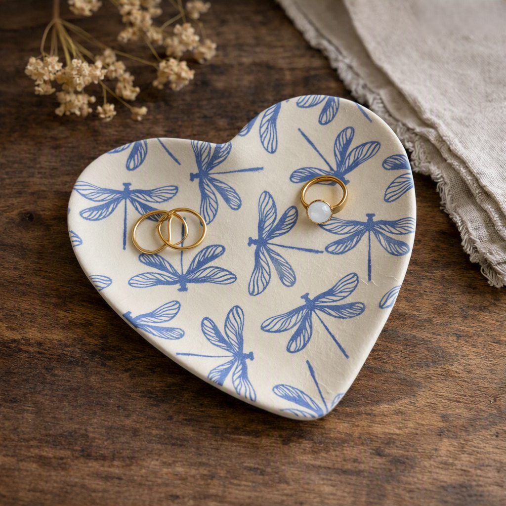 A heart-shaped ceramic dish with a blue dragonfly pattern, styled on a dark wooden table with dried flowers and a linen cloth. Two gold rings are placed on the dish, one with a white stone.