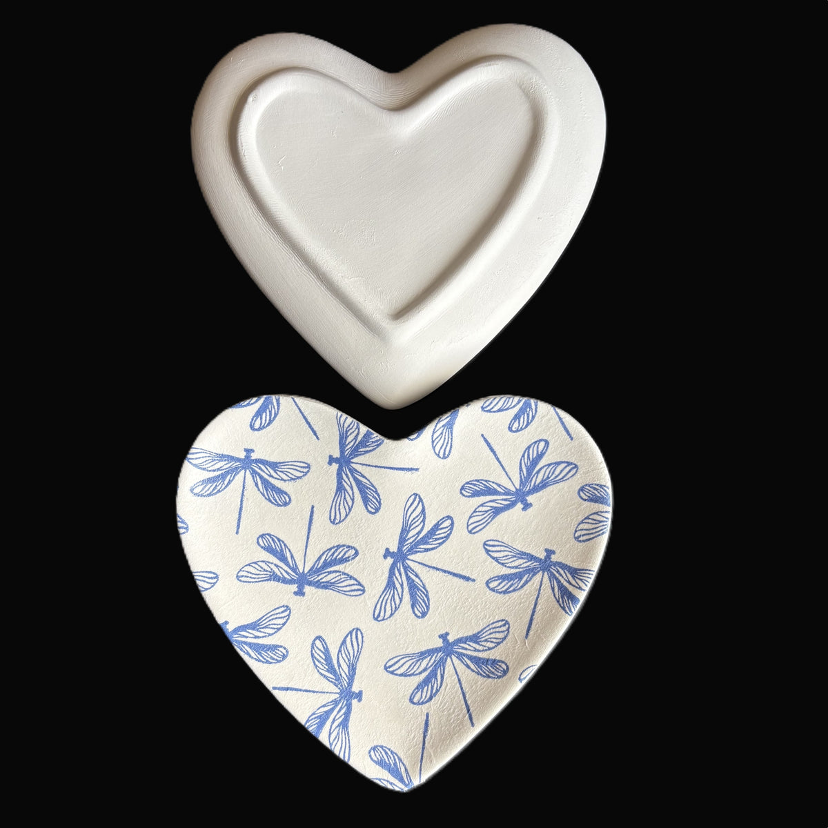 Two heart-shaped ceramic Trinket Tray shown against a black background: one plain off-white on top, and one white with a blue dragonfly pattern on the bottom.