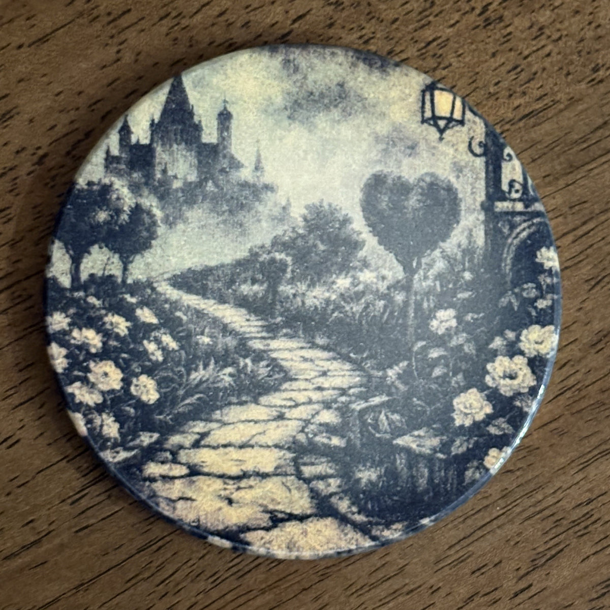 A round coaster featuring a vintage-style blue and cream illustration of a stone path leading to a castle, surrounded by flowers and trees, displayed on a wooden surface.