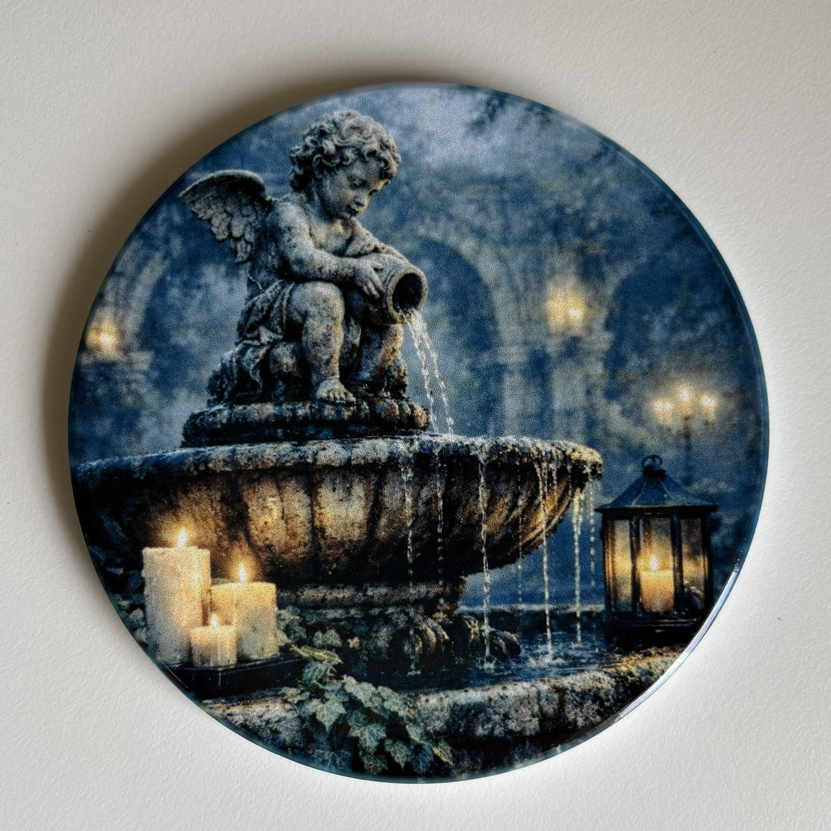 A round decorative coaster featuring a printed scene of a stone fountain with a winged cherub statue, water cascading down, candles, and a lantern in a blue-toned architectural setting.