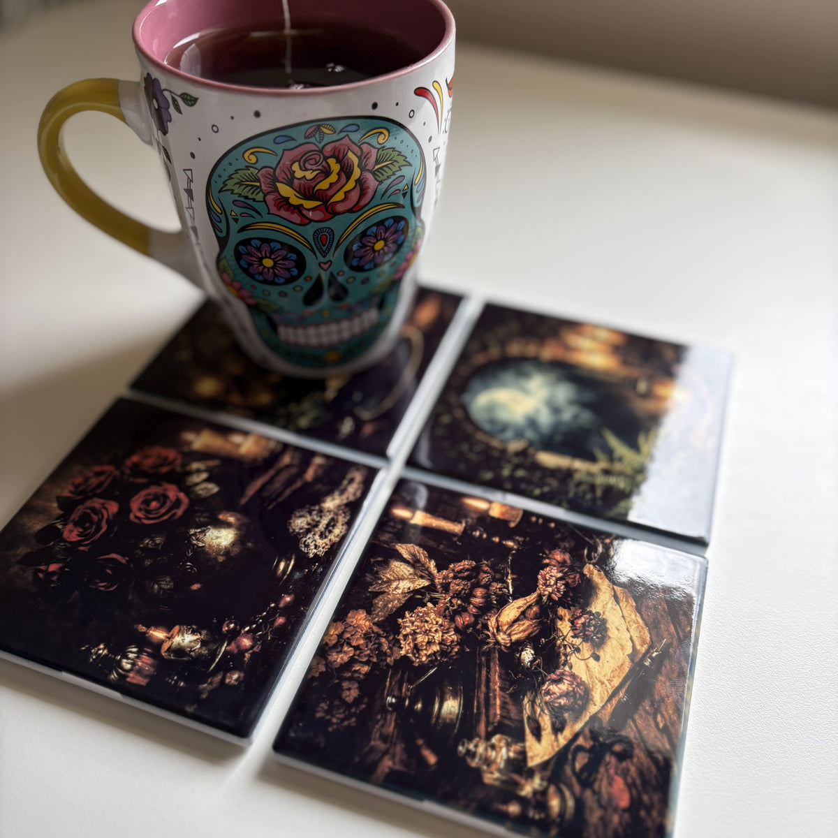 Four square coasters with dark, moody floral and vintage illustrations arranged on a white tabletop, with a colorful ceramic mug placed on top.