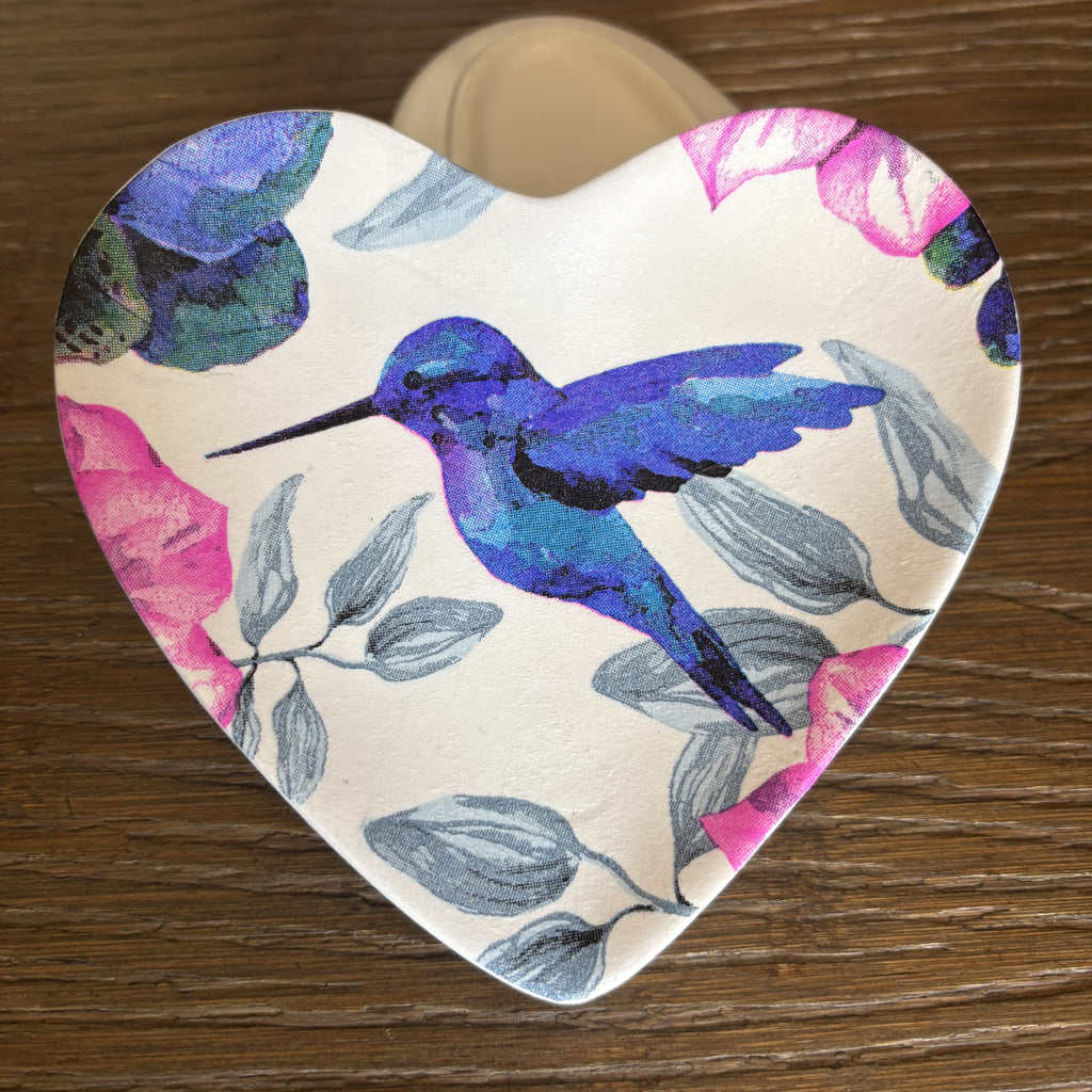 A heart-shaped ceramic trinket tray featuring a vibrant blue and purple hummingbird and pink floral design on a cream background, set on a dark wood surface.