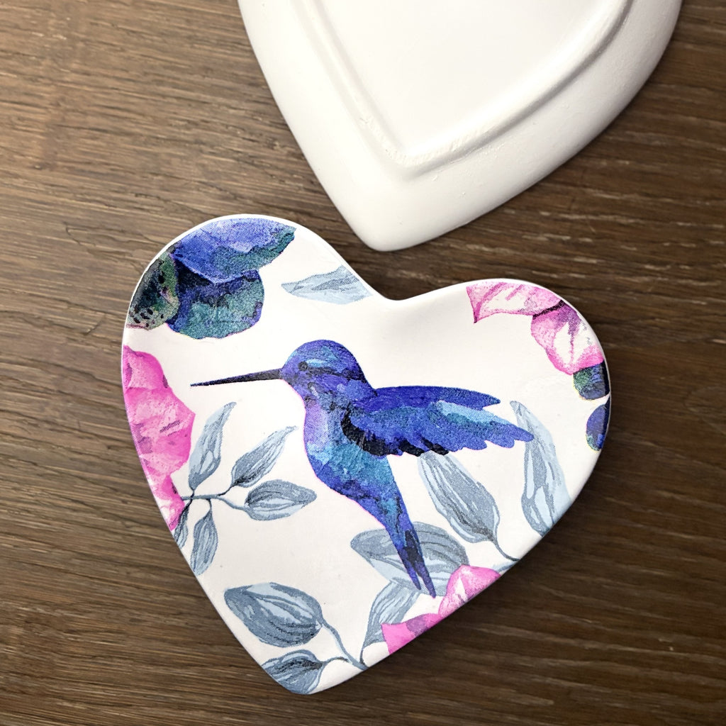 A heart-shaped ceramic trinket tray featuring a watercolor design of a blue hummingbird and pink flowers on a white background, placed on a dark wood surface next to a white ceramic object.