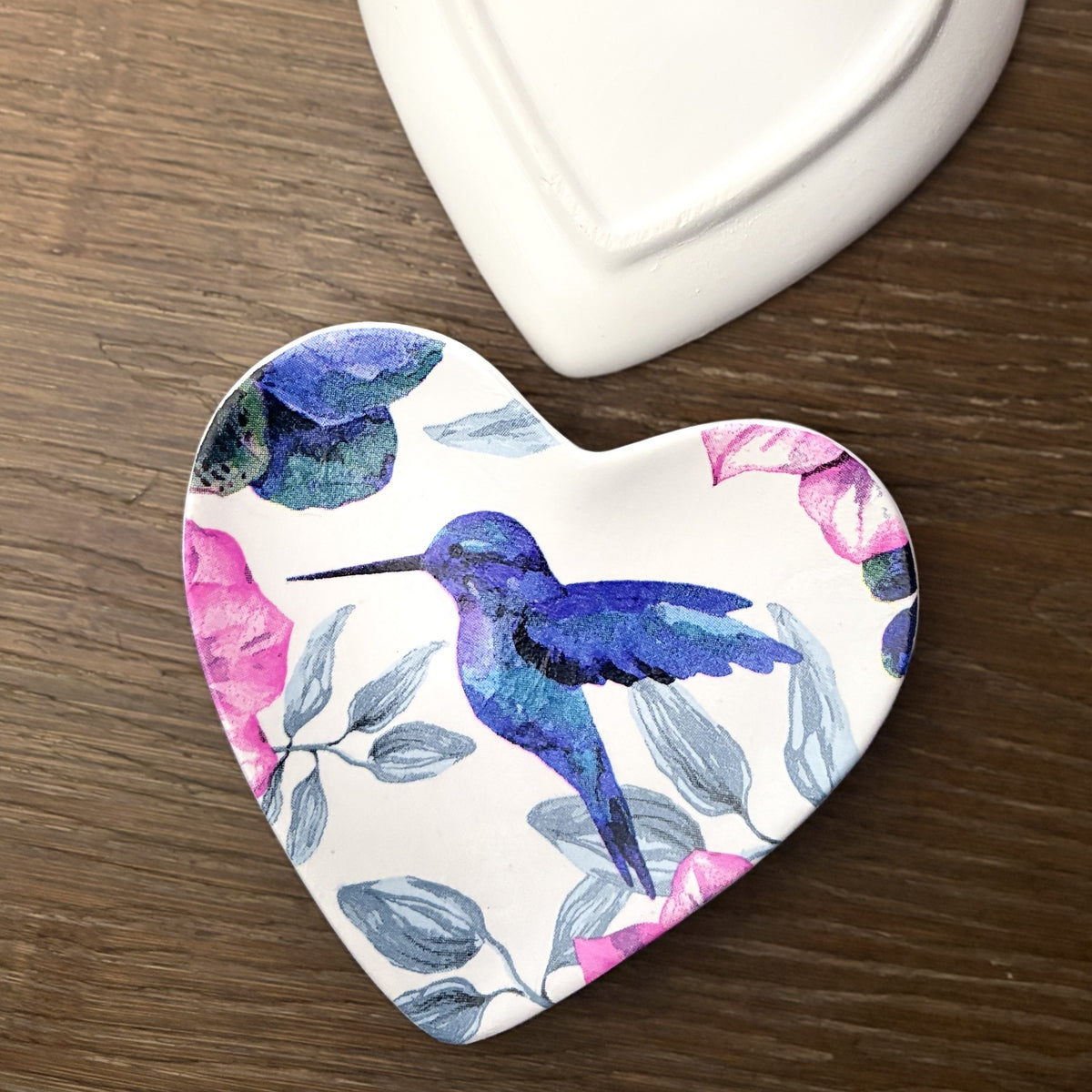 A heart-shaped ceramic trinket tray featuring a watercolor design of a blue hummingbird and pink flowers on a white background, placed on a dark wood surface next to a white ceramic object.