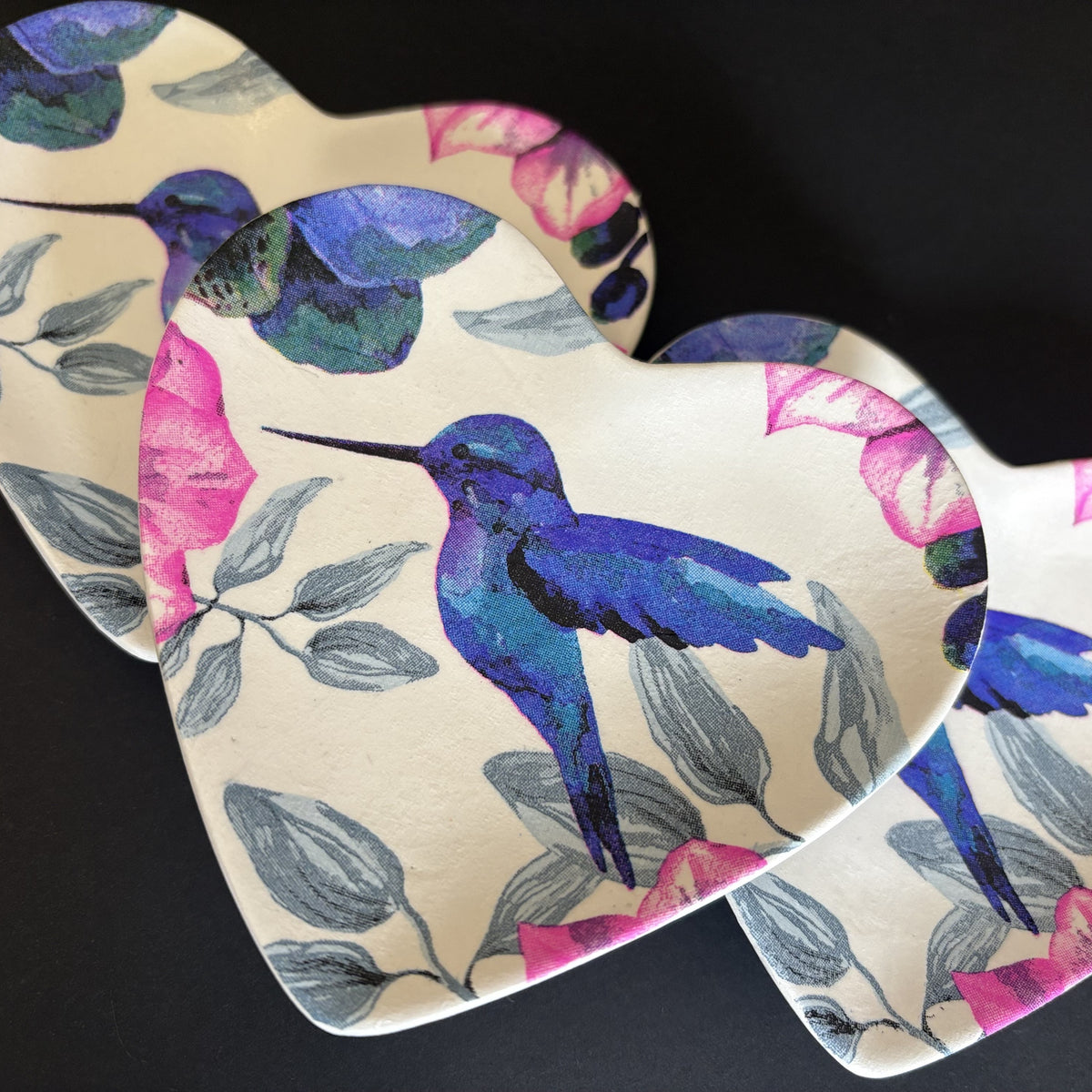 Three heart-shaped trinket trays with a vibrant hummingbird and floral pattern, displayed on a dark background.
