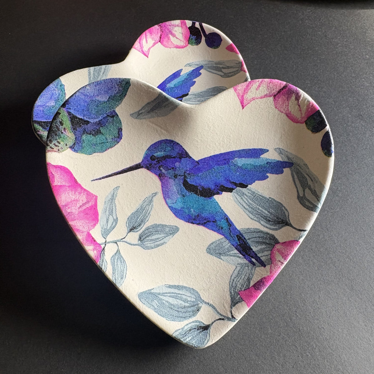 A heart-shaped ceramic trinket tray with a colorful print of a blue and purple hummingbird, pink flowers, and gray leaves on a cream background, set against a dark gray surface.