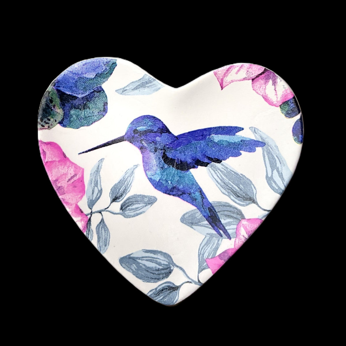 A heart-shaped ceramic trinket tray featuring a watercolor-style design of a blue hummingbird, pink flowers, and gray leaves on a white background, set against a solid black backdrop.