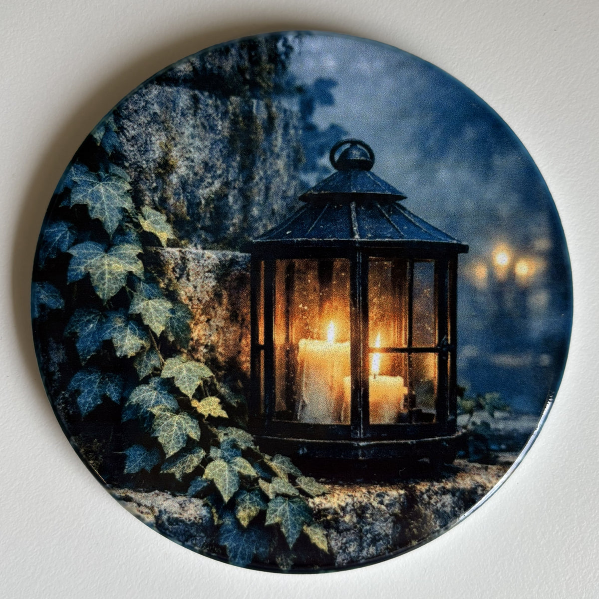 A round decorative coaster featuring a printed scene of a black lantern with glowing candles against a stone wall and ivy, photographed on a light neutral background.