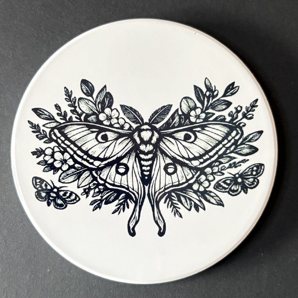Round white coaster with black butterfly and floral design on a dark background