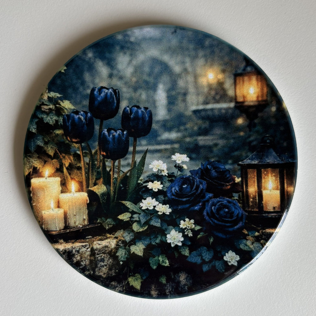 A round decorative coaster featuring a gothic floral scene with dark blue tulips and roses, white flowers, candles, and lanterns against a blurred stone background, photographed against a plain light wall.