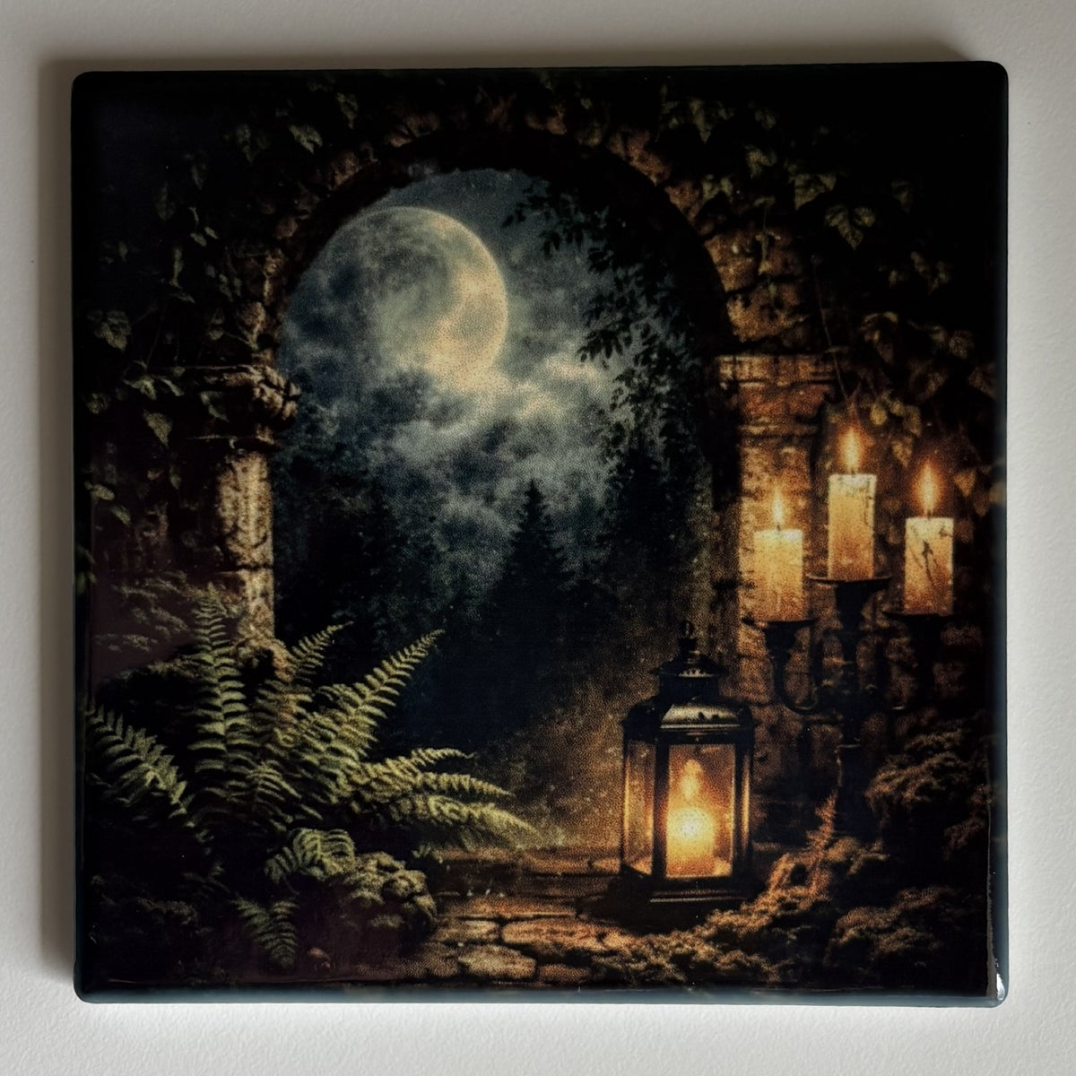 A square coaster featuring a dark fantasy night scene with an arched stone opening, a full moon, ferns, candles, and a lantern, shown against a plain light background.