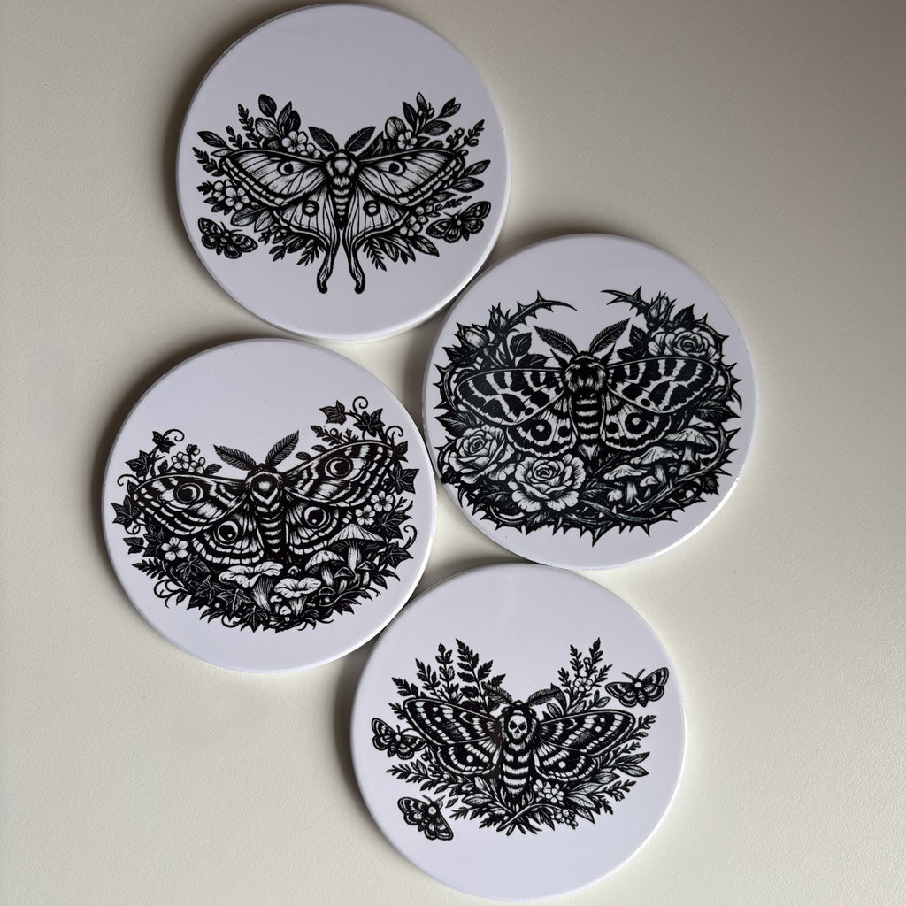 Four round white coasters with black moths and floral illustrations, arranged on a light beige background.
