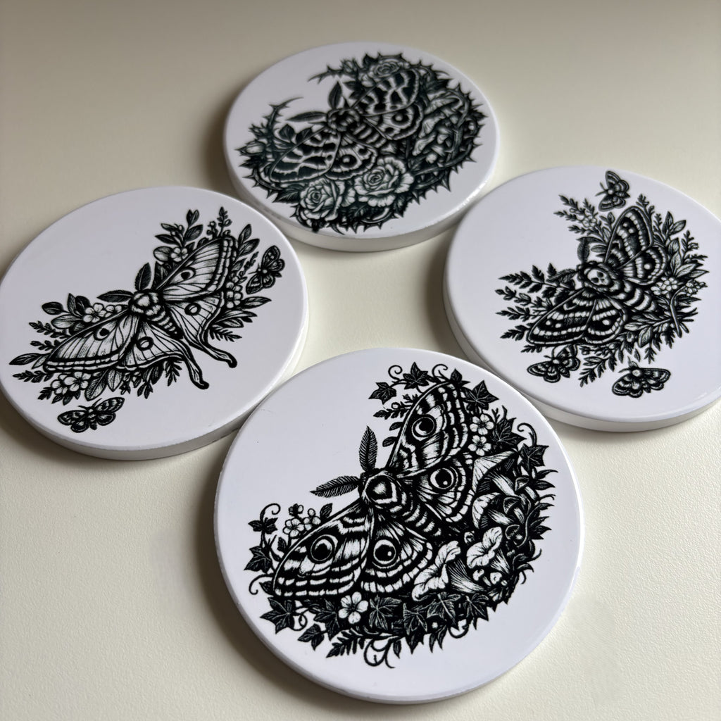 Four round white coasters with black floral and moth illustrations, arranged on a light beige surface.