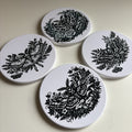 Four round white coasters with black floral and moth illustrations, arranged on a light beige surface.